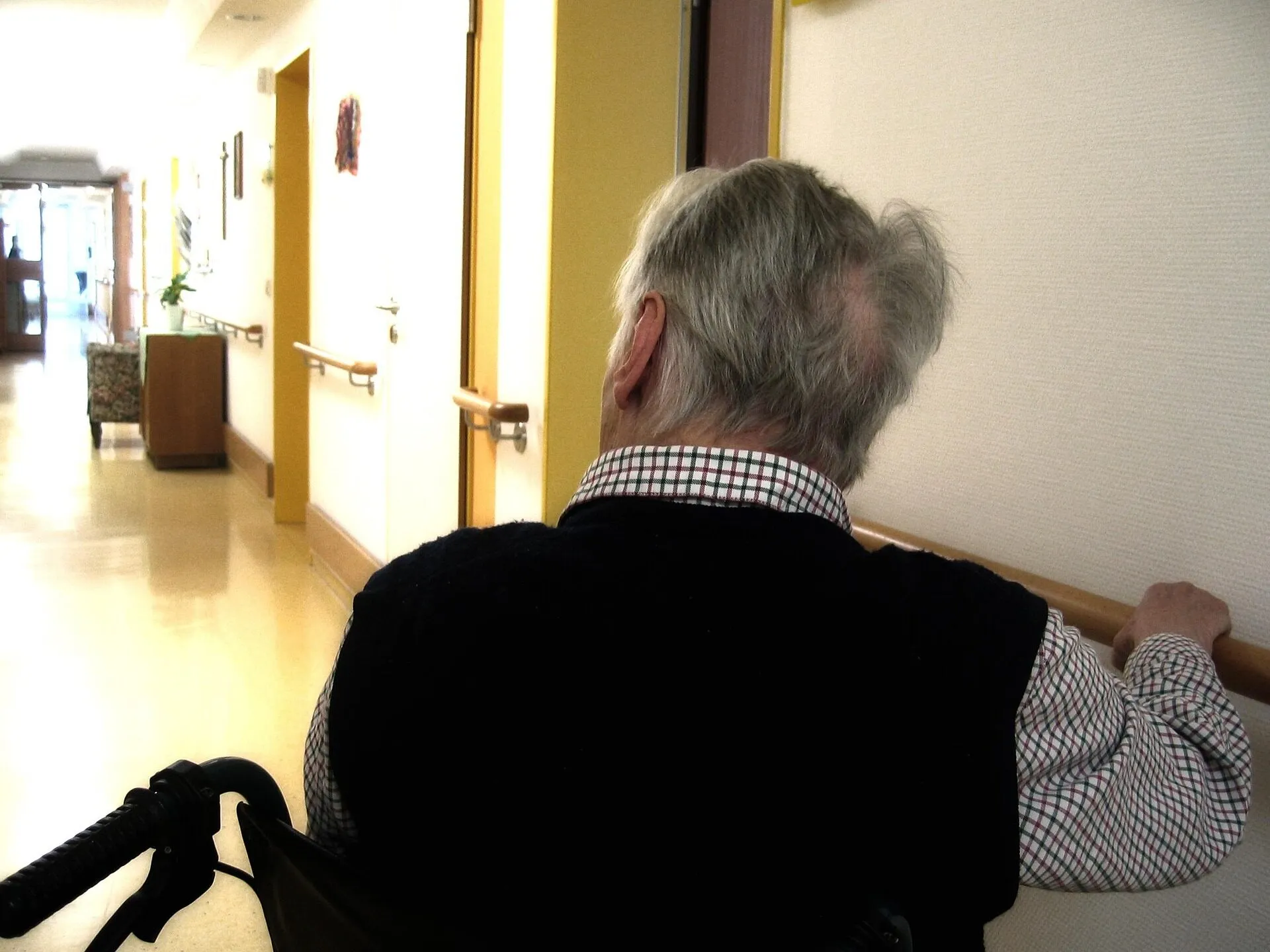 Résident âgé en fauteuil roulant, de dos, tenant la main courante d’un corridor de résidence